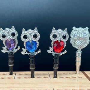 May include: Five silver owl-shaped phone charms with jeweled eyes and different colored gem accents. The owls are perched on a branch-like design and have a silver post for attaching to a phone case.