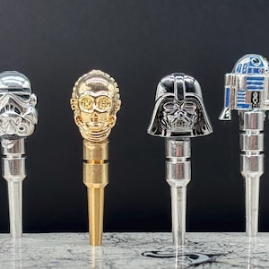 May include: Four metal bottle stoppers featuring iconic Star Wars characters: a Stormtrooper, C-3PO, Darth Vader, and R2-D2. The stoppers have detailed character designs and are silver, gold, and blue. The stoppers are displayed against a dark background.