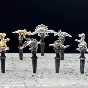 May include: A collection of decorative bottle stoppers featuring dinosaur designs. The stoppers are in gold and silver tones, with black bases. The dinosaurs include a T-Rex, Stegosaurus, and other prehistoric creatures.