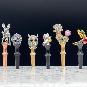 May include: A collection of decorative bottle stoppers with animal and floral designs. Each stopper features a different jeweled design, including a rabbit, owl, mouse, poodle, and hummingbird. The stoppers have a variety of colors and are set against a dark background.