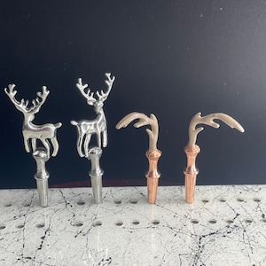May include: Four decorative metal deer finials in silver and copper finishes. The deer are standing on a white surface with black speckles.