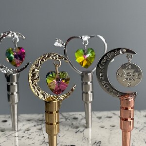 May include: Four decorative bottle stoppers with different designs. The first is silver with a crescent moon and a pink heart. The second is gold with a crescent moon and a rainbow heart. The third is silver with a heart and a green heart. The fourth is silver with a crescent moon and a compass.