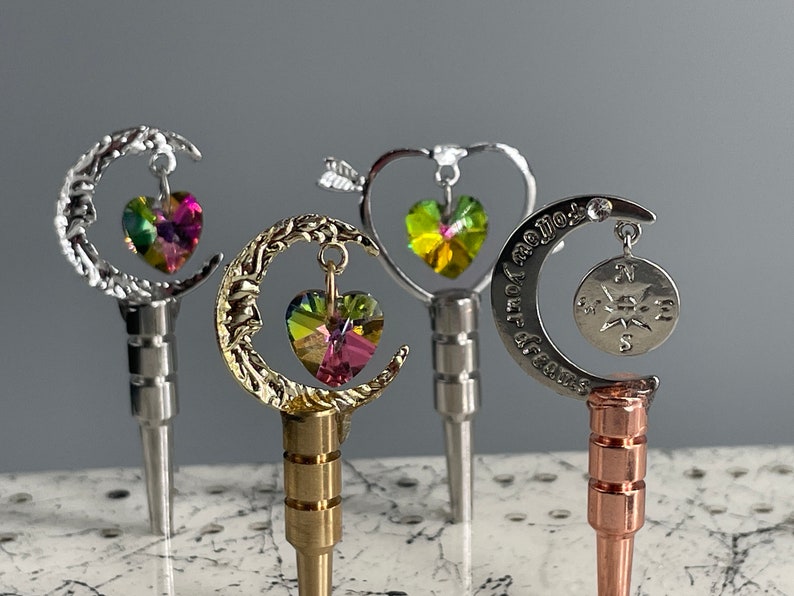 May include: Four decorative bottle stoppers with different designs. The first is silver with a crescent moon and a pink heart. The second is gold with a crescent moon and a rainbow heart. The third is silver with a heart and a green heart. The fourth is silver with a crescent moon and a compass.