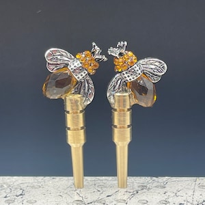 May include: Two gold metal bee-shaped bottle stoppers with clear amber glass accents and rhinestone details.