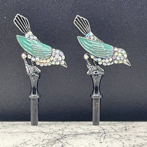 May include: Two decorative metal bird finials with green enamel and iridescent rhinestones. The birds are perched on black metal posts.