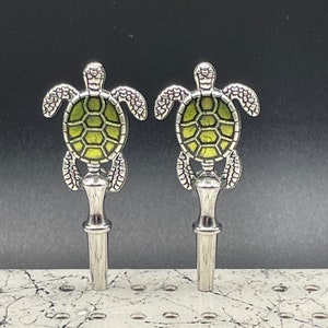 May include: Two silver bottle stoppers with a green turtle design. The turtles have a detailed shell and are facing forward.