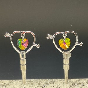 May include: Two silver keychains with a heart-shaped design and an arrow piercing through the heart. Each heart features a multi-colored crystal in the center.