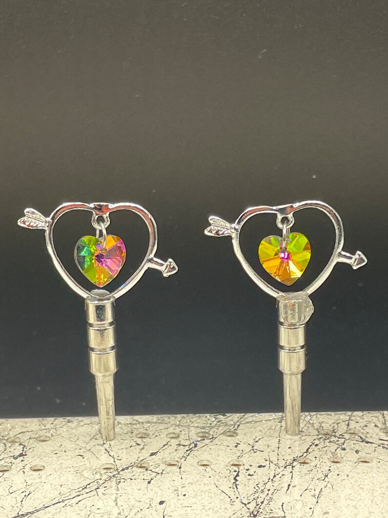 May include: Two silver keychains with a heart-shaped design and an arrow piercing through the heart. Each heart features a multi-colored crystal in the center.