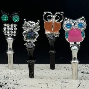 May include: Four owl-shaped bottle stoppers with different designs. The stoppers are made of metal and have a silver finish. The owls are decorated with rhinestones, enamel, and other embellishments.