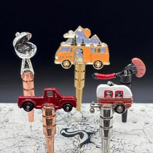 May include: Five miniature metal figurines on metal rods. The figurines are a red pickup truck, a white camper, a grill, a yellow camper van with a mountain scene, and a red wrench with a sausage.