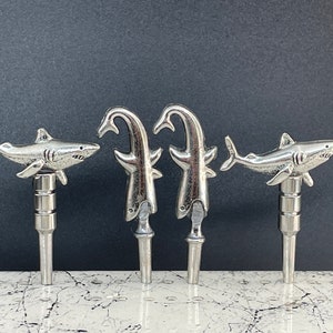 May include: Four silver bottle stoppers shaped like sharks. Two stoppers are shaped like a shark fin and two are shaped like a shark with an open mouth.