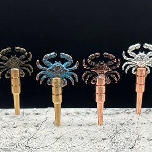 May include: Five metal bottle stoppers with crab designs. The crabs are in different colors: black, blue, copper, and silver.