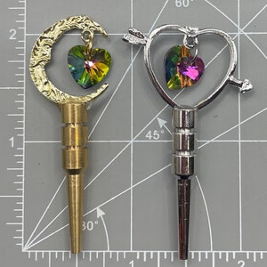 May include: Two decorative bottle stoppers with a heart-shaped crystal charm. One stopper is gold-toned and shaped like a crescent moon. The other stopper is silver-toned and shaped like a heart with an arrow through it.