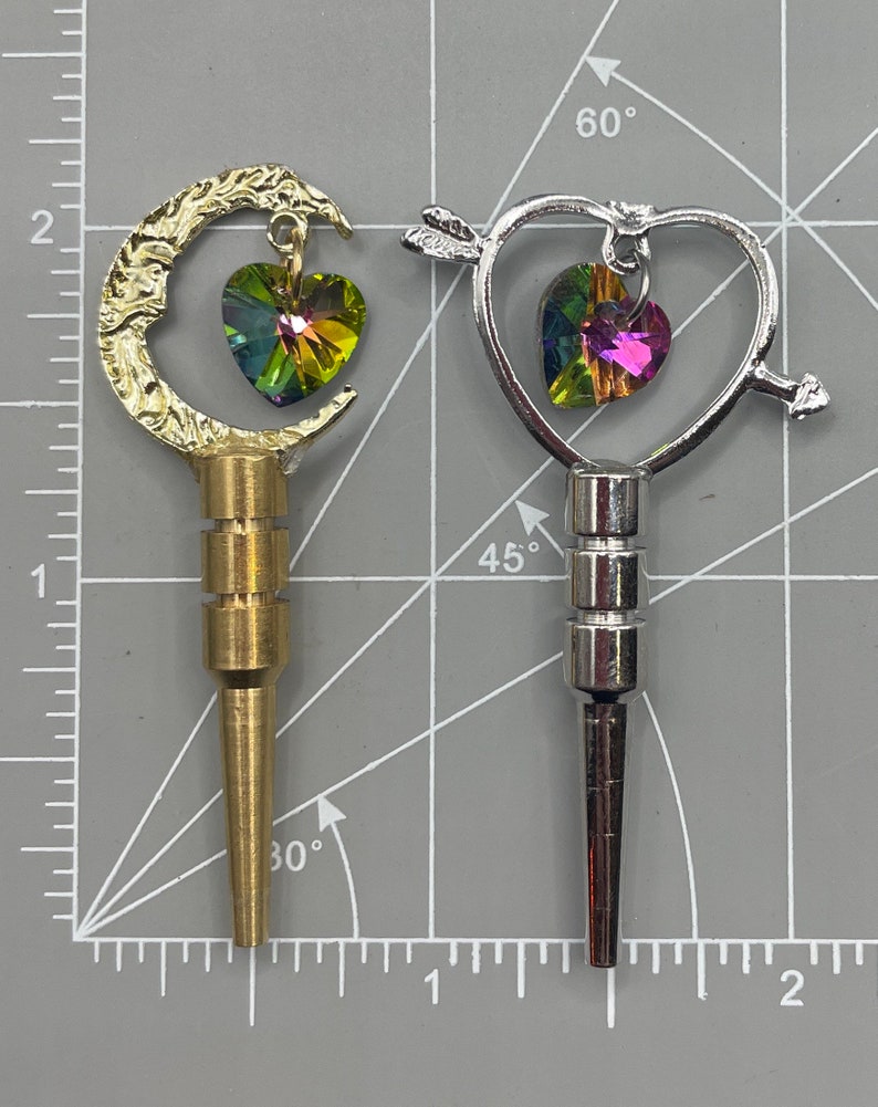 May include: Two decorative bottle stoppers with a heart-shaped crystal charm. One stopper is gold-toned and shaped like a crescent moon. The other stopper is silver-toned and shaped like a heart with an arrow through it.
