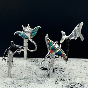 May include: Four silver and abalone shell sea creature bottle stoppers. The stoppers are shaped like a manta ray, a whale, a dolphin, and a stingray.