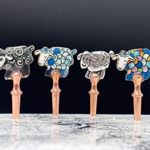 May include: Four decorative bottle stoppers shaped like sheep. Each stopper is made of metal and features a different design. The stoppers are all in a row on a marble surface.