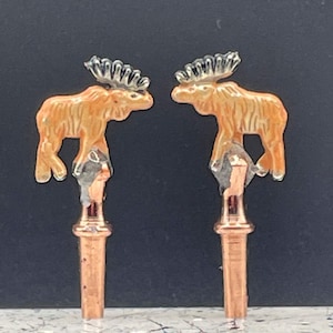 May include: Two copper bottle stoppers with moose figurines on top. The moose are orange with black antlers and are facing each other.