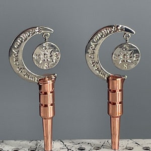 May include: Two silver crescent moon bottle stoppers with a dangling star charm. The stoppers are attached to a copper colored base.