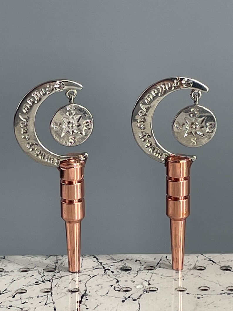 May include: Two silver crescent moon bottle stoppers with a dangling star charm. The stoppers are attached to a copper colored base.