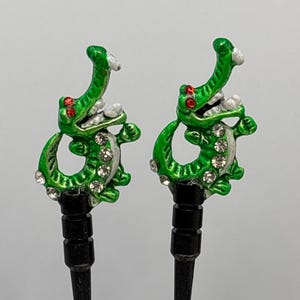 May include: A pair of green alligator golf tees with red eyes and rhinestones. The tees are black and have a pointed tip.