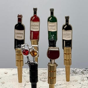May include: A collection of decorative wine bottle stoppers. The stoppers feature colorful enamel designs of wine bottles and glasses. One stopper has the text "LAFITE 1982". The stoppers have gold and black bases.