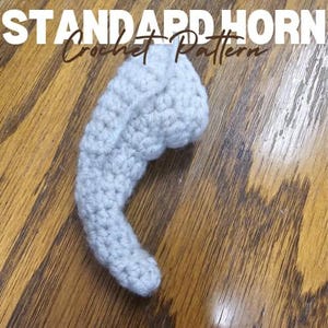 Peut inclure: Une corne crochetée gris clair, posée sur une surface en bois. La corne est faite avec un motif de point serré et a une forme incurvée. Le texte "STANDARD HORN Crochet Pattern" est en haut de l'image.
