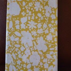 Può includere: Un libro rettangolare con una copertina a motivi floreali gialli. La copertina presenta fiori e foglie bianche su sfondo giallo. Il libro è posizionato verticalmente, con il dorso rivolto verso l'osservatore.