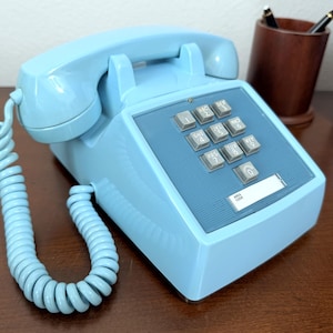 May include: A vintage light blue telephone with a push-button keypad. The phone has a coiled handset cord and a rectangular base. The keypad has gray buttons and a label area. The phone is on a wooden surface.