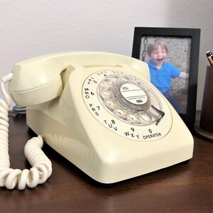 May include: A vintage rotary dial phone with a cream-colored body and a black rotary dial. The phone has a coiled cord and is sitting on a wooden surface.