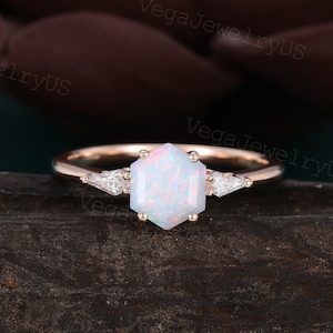 Könnte beinhalten: Ein Verlobungsring aus Roségold mit einem sechseckigen Opal-Edelstein und zwei kleineren Diamanten. Der Ring liegt auf einer braunen Holzoberfläche.