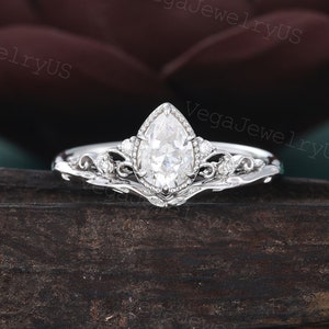 May include: A pear-shaped diamond engagement ring with a delicate, ornate band. The ring is set in white metal and features a pear-shaped diamond in the centre, surrounded by smaller diamonds.
