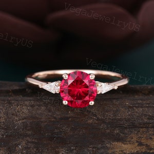 May include: A rose gold engagement ring featuring a large, round, red ruby gemstone surrounded by two smaller, triangular, white diamonds.