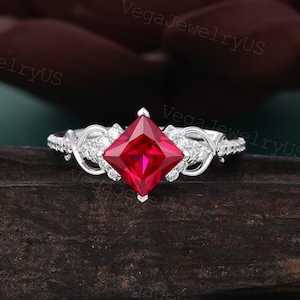 May include: A silver ring with a square-shaped ruby gemstone surrounded by smaller diamonds. The ring has a delicate, twisted band.