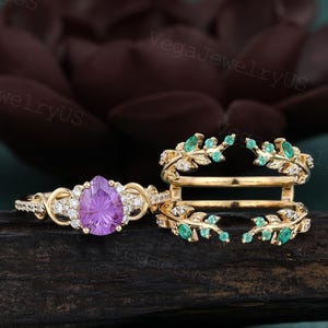 May include: A gold ring set featuring a pear-shaped purple gemstone and a ring guard. The engagement ring has a halo of small diamonds. The ring guard is adorned with green gemstones and small diamonds, with a leaf design.
