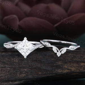 Puede incluir: Dos anillos a juego. El anillo de compromiso presenta un diamante talla princesa y una banda retorcida con pequeñas piedras de acento. La alianza tiene un diseño curvo con detalles en forma de hoja y pequeños diamantes. Ambos anillos son plateados.