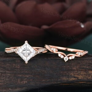 May include: A rose gold engagement ring set. The engagement ring features a princess-cut diamond and a band with small accent stones. The wedding band has a curved design with small accent stones. The rings are displayed on a dark wooden surface.
