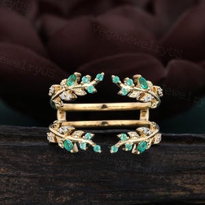 May include: A gold-colored ring with a leaf design, featuring small green and clear gemstones. The ring has two bands that curve around each other, creating a unique and elegant look. The gemstones are set in a delicate pattern.