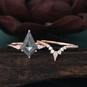 May include: A set of two rose gold engagement rings with a black diamond center stone and a delicate diamond band. The center stone is a kite-shaped black diamond with a gray hue. The band is a delicate pave setting with small round diamonds.