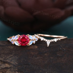 May include: A rose gold engagement ring set with a red ruby center stone and a halo of moonstone accents. The matching wedding band features a delicate, curved design with small diamonds.