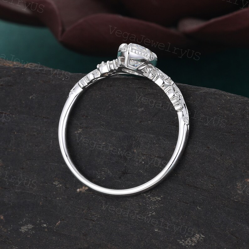 May include: A silver engagement ring with a round centre stone and smaller stones on the band. The ring is set on a dark green surface.