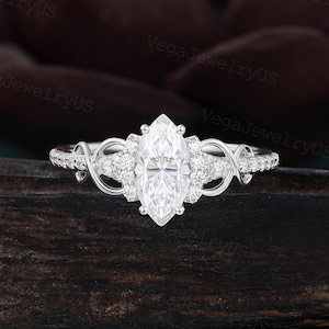 May include: A silver-toned ring featuring a central marquise-cut clear stone flanked by small round stones. The band includes intricate swirling metalwork and additional small round stones along the sides, creating a detailed and ornate appearance.