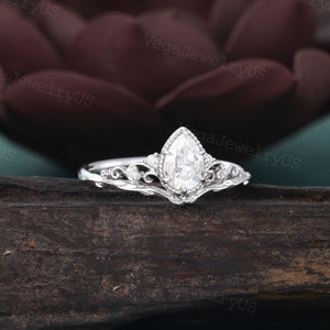 May include: A pear-shaped diamond engagement ring with a delicate, ornate band. The ring is set in white gold and features a floral design.