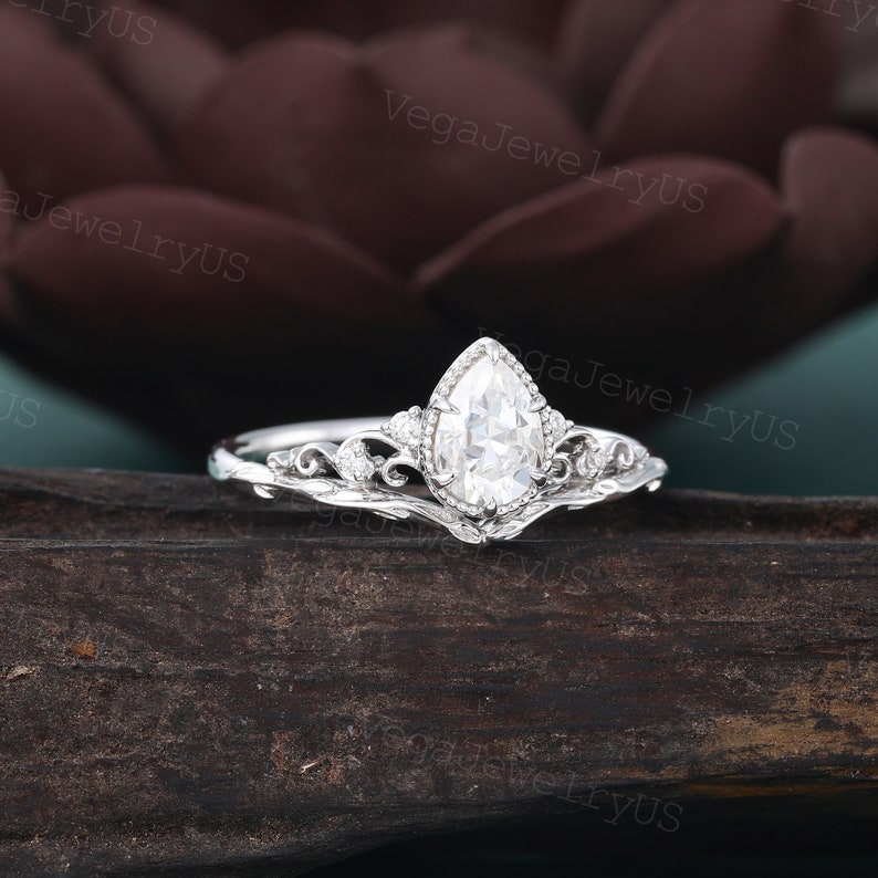May include: A pear-shaped diamond engagement ring with a delicate, ornate band. The ring is set in white gold and features a floral design.