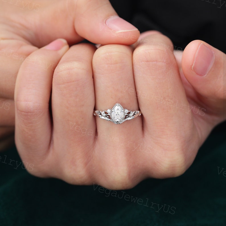 May include: A pear-shaped diamond engagement ring with a delicate crown-like setting. The ring is set in white gold and features a sparkling diamond.
