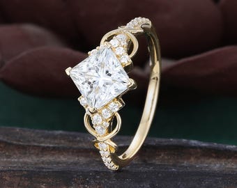 May include: A gold ring featuring a large, square-cut, clear center stone set in a four-prong mount. The band is accented with smaller, round, clear stones along the shoulders and includes decorative, intertwined gold loops on both sides.