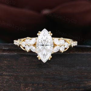 Op de afbeelding: Een verlovingsring met een marquise-geslepen diamant en een gouden band. De ring heeft een grote centrale diamant en kleinere diamanten en bloemaccenten op de band. De ring is geplaatst op een donkere houten ondergrond.