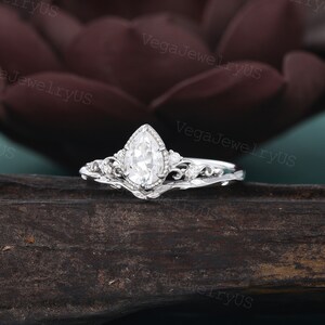 May include: A pear-shaped diamond engagement ring set in a delicate, ornate silver band with a matching wedding band. The ring features a pear-shaped diamond with a halo of smaller diamonds and intricate floral details.