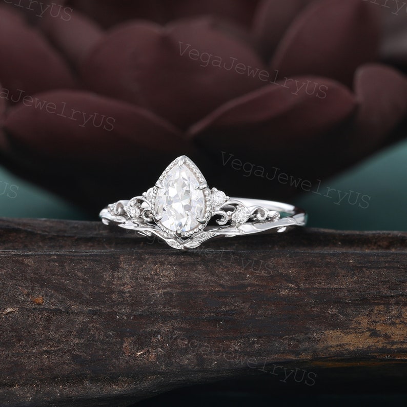 May include: A pear-shaped diamond engagement ring set in a delicate, ornate silver band with a matching wedding band. The ring features a pear-shaped diamond with a halo of smaller diamonds and intricate floral details.