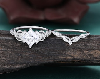 May include: A silver-toned engagement ring set featuring a princess-cut center stone flanked by intricate scrollwork and small round accent stones, paired with a matching curved wedding band adorned with marquise and round-cut stones.