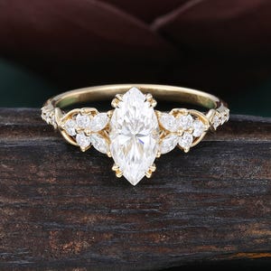 May include: A gold ring featuring a large marquise-cut center stone held by prongs. The band is adorned with smaller marquise and round-cut stones arranged in floral-like clusters on both sides of the center stone, extending along the shank.